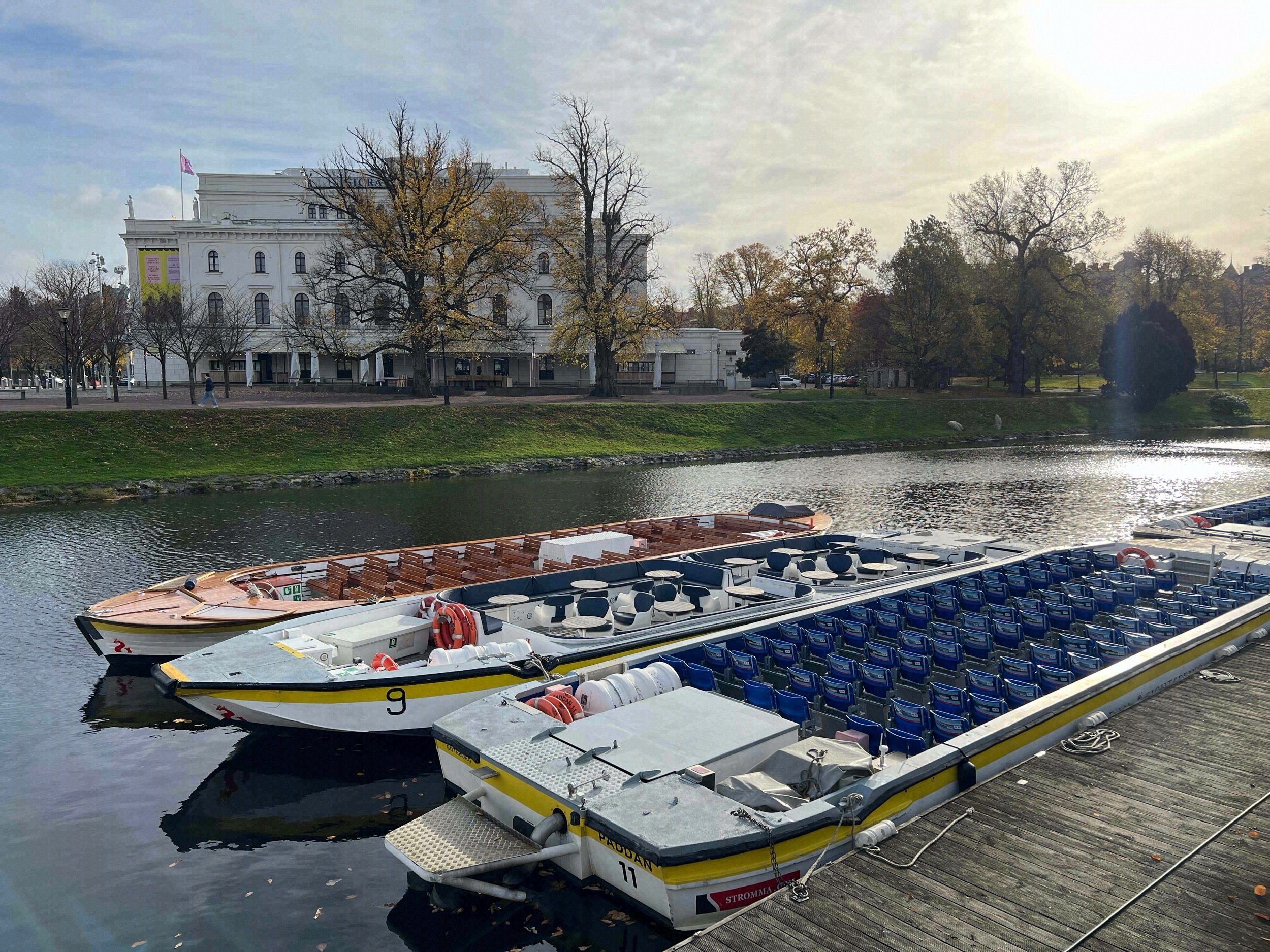 Paddan-Kanalboote liegen an einem Steg, im Hintergrund spiegelt sich der Stora Teatern im Parkweiher des Trädgårdsföreningen.