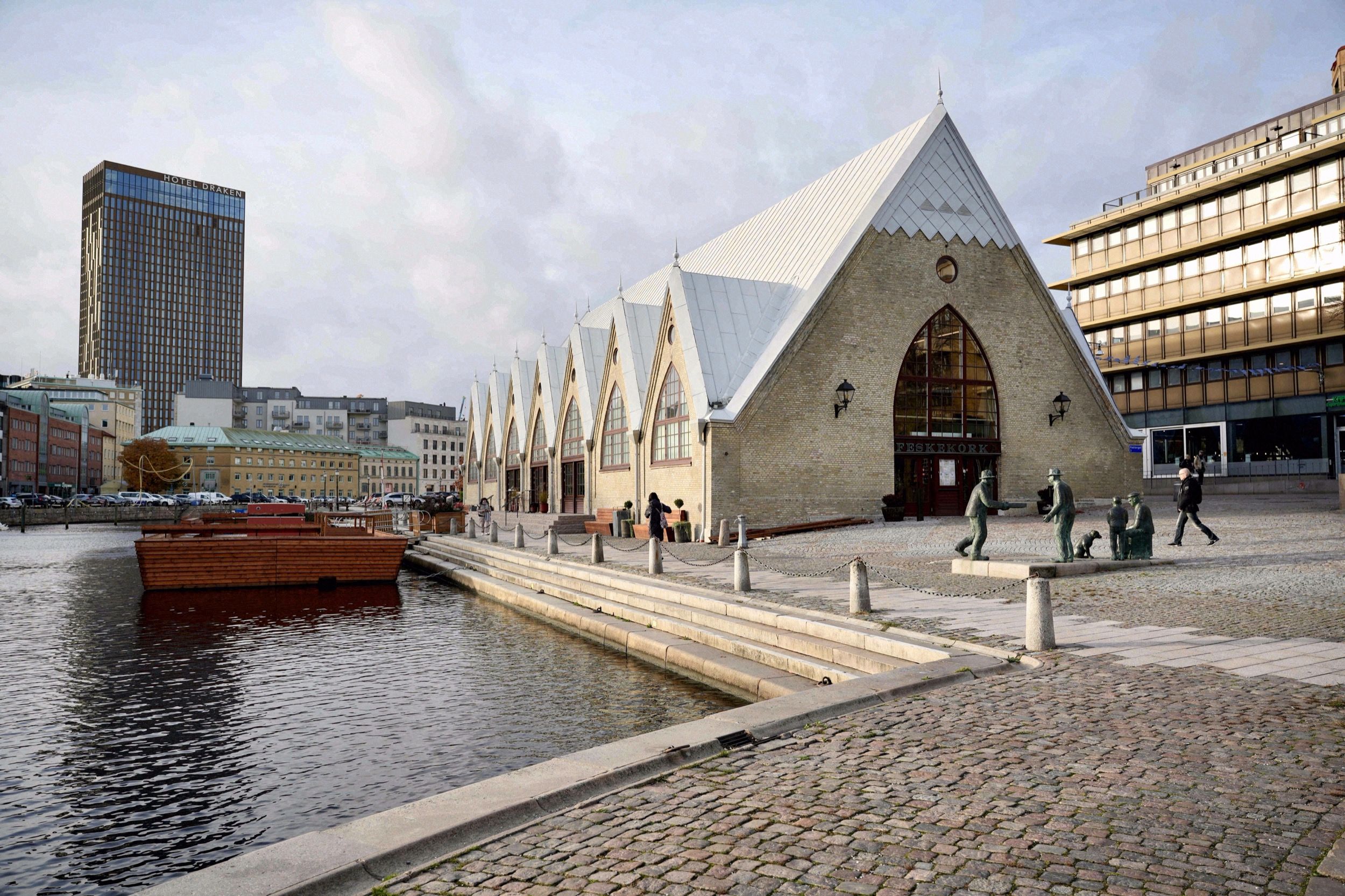 Frontansicht der Feskekôrka vom Kanal aus mit Bronzeskulpturen auf dem Pflasterplatz und dem Innenstadt-Panorama im Hintergrund.